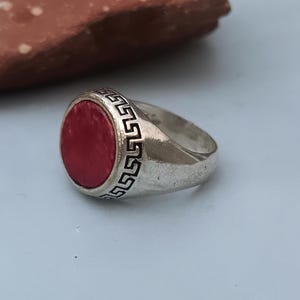 Puede incluir: Un anillo de tono plateado con una incrustación de piedra roja circular y plana en el centro. La banda de metal está decorada con un patrón de greca griega relleno de negro que rodea la piedra roja, creando un contraste con la superficie.