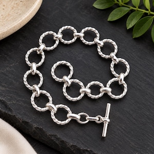 Può includere: Un bracciale color argento composto da una serie di maglie circolari interconnesse con una texture a corda ritorta. Il bracciale presenta un meccanismo di chiusura a T composto da un anello circolare e da una barra dritta.