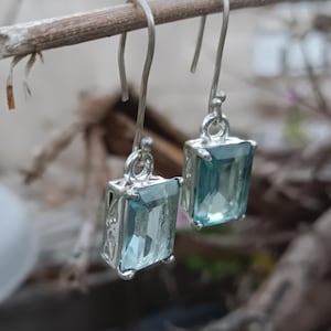 May include: A pair of silver drop earrings featuring rectangular, light blue faceted gemstones set in ornate metal frames. The earrings hang from simple silver hooks, showcasing a clear, polished finish on the stones and intricate metalwork.