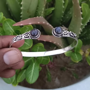 Könnte beinhalten: Ein silberner Armreif in offenem Design mit zwei runden, dunkelvioletten Cabochon-Steinen an den Enden. Die Metallverarbeitung um die Steine herum zeigt komplizierte Schnörkelmuster und kleine perlenartige Details an den Enden des Armreifs.