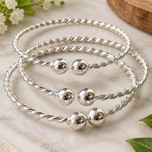 Può includere: Tre bracciali rigidi in metallo color argento con una texture intrecciata simile a una corda. Ogni bracciale aperto è rifinito con una perla sferica liscia e lucida su entrambe le estremità. I bracciali sono disposti in una formazione sovrapposta, mettendo in risalto la superficie riflettente del metallo e il motivo a spirale costante lungo le fasce circolari.