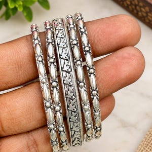Può includere: Un set di cinque bracciali rigidi in metallo color argento con finitura lucida. Quattro bracciali presentano un motivo ripetitivo di piccole sfere e forme ovali allungate, mentre il bracciale centrale mostra un dettagliato motivo floreale in rilievo.
