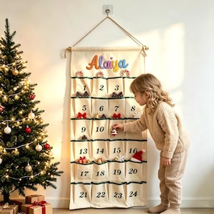 Pode incluir: Um calendário do advento de tecido cor de creme para pendurar na parede com vinte e quatro bolsos numerados de um a vinte e quatro em fonte verde escura. O nome Alaiya está bordado em letras coloridas no topo com pequenos enfeites de Natal.