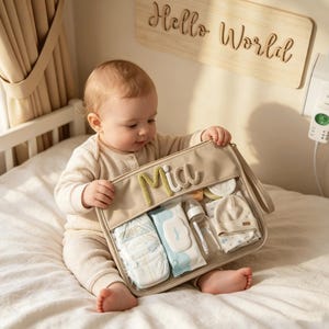 May include: A beige rectangular organizer bag with a clear front panel, featuring the name Mia in textured green and white letters. The bag contains a stack of diapers, a pack of baby wipes, a clear plastic baby bottle, and a folded beige baby hat.
