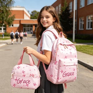 Könnte beinhalten: Ein hellrosa Rucksack und eine passende Lunchtasche, beide mit einem Muster aus kleinen dunkelrosa Schleifen und Punkten. Jeder Artikel ist mit dem Namen Grace in einer dunkelrosa Schreibschrift personalisiert. Der Rucksack hat verstellbare Träger.
