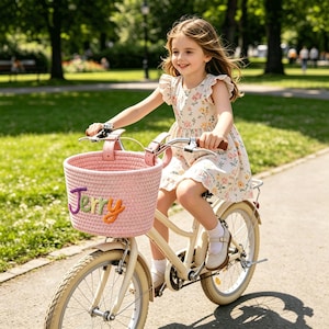 Puede incluir: Una cesta de bicicleta de cuerda tejida de color rosa claro con el nombre Jerry bordado en letras de hilo de colores. La cesta está sujeta al manillar de una bicicleta de color crema, con un vestido de flores y zapatos blancos visibles.