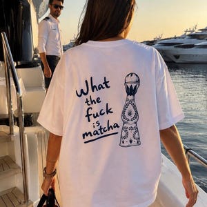 Könnte beinhalten: Ein weißes Kurzarm-T-Shirt mit einer dunkelblauen Grafik auf der Rückseite. Das Design zeigt eine stilisierte Abbildung einer Tequila-Flasche neben dem Text "What the fuck is matcha" in einer lockeren, handschriftlichen Schriftart.
