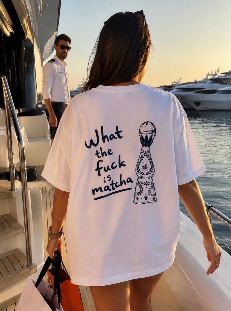 May include: A white short-sleeved t-shirt featuring a dark blue graphic on the back. The design includes a stylised illustration of a tequila bottle next to the text "What the fuck is matcha" written in a casual, handwritten-style font.