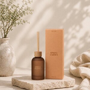 Puede incluir: Un frasco difusor de varillas de vidrio esmerilado marrón con tapa de madera y tres varillas, acompañado de una caja de cartón rectangular a juego. Tanto el frasco como la caja presentan el texto SPRING GARDEN en una fuente sans-serif marrón.