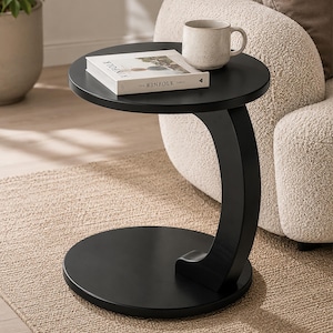 Può includere: Un tavolino nero con piano e base rotondi, collegati da un supporto curvo. Un libro intitolato "The Kinfolk Table" e una tazza beige sono sul tavolo. Il tavolo è su un tappeto beige, vicino a una poltrona color crema.