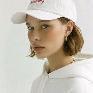 May include: A white baseball cap featuring the word Invoicing embroidered in red script font on the front panel. The cap has a structured crown, a curved brim, and a matching white fabric construction, providing a clean and simple aesthetic.