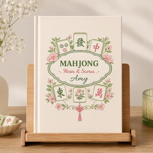 Puede incluir: Un libro de tapa dura color crema titulado Mahjong Notes & Scores con el nombre Amy impreso en la portada. La cubierta presenta ilustraciones de fichas de mahjong, flores de cerezo rosas, hojas verdes y una borla decorativa roja.