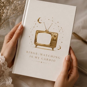 Op de afbeelding: Een crèmekleurig notitieboek met een gouden illustratie van een vintage televisie, sterrenbeelden en de tekst "Binge-Watching Is My Cardio". Het notitieboek wordt door een persoon vastgehouden.