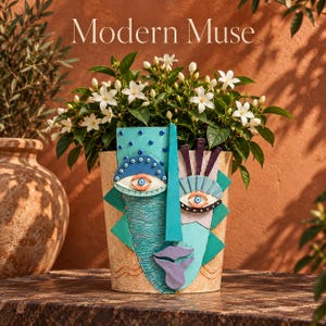 May include: A beige ceramic planter featuring a colourful abstract face design with teal, blue, and purple geometric shapes. The planter holds a green plant with white flowers. The text Modern Muse is printed in white above the planter.