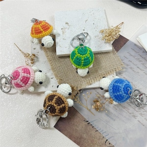 May include: Five small crochet turtle keychains with white heads and shells in orange, green, pink, brown, and blue. Each turtle features a silver metal keyring attached to the top of the shell, showcasing detailed yarn stitching and round black eyes.