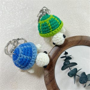 May include: Two crocheted turtle keychains with silver metal rings. One turtle features a bright green shell with a white head and black bead eyes, while the other has a vibrant blue shell with a matching white head and black bead eyes.