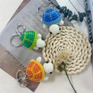May include: Three crochet turtle keychains featuring shells in blue, green, and orange with cream-colored heads and black bead eyes. Each turtle is attached to a silver-toned metal split ring. The turtles are arranged on a surface.