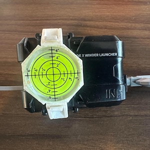 Könnte beinhalten: Ein schwarz-weißer Spielzeugwerfer mit einer Wasserwaage und einer Zugschnur. Der Werfer hat eine kreisförmige Wasserwaage mit Gradmarkierungen und dem Text "DE X WINDER LAUNCHER". Der Werfer befindet sich auf einer Holzoberfläche.