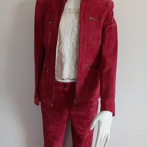 May include: A deep red suede jacket and matching trousers set worn over a cream-colored top with a lace panel. The jacket features a high collar, a central zipper, and two horizontal zippered pockets on the chest and waist area.