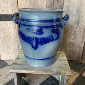 Peut inclure: Un pot en grès bleu et gris avec deux poignées et des motifs décoratifs bleu foncé. Le pot est posé sur un petit tabouret en bois vieilli. L'arrière-plan est un mur en bois.