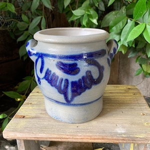 Può includere: Un vaso in gres grigio chiaro con due manici laterali e un prominente motivo floreale dipinto a mano in blu cobalto che circonda la parte superiore. Il contenitore presenta un'ampia apertura circolare con bordo spesso e finitura smaltata.