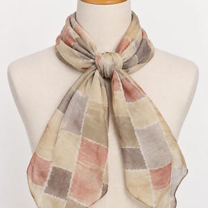 May include: A lightweight scarf featuring a repeating patchwork pattern of muted squares in shades of dusty rose, beige, taupe, and soft gray. The fabric has a semi-sheer, smooth texture and is tied in a simple knot at the center of the neck.