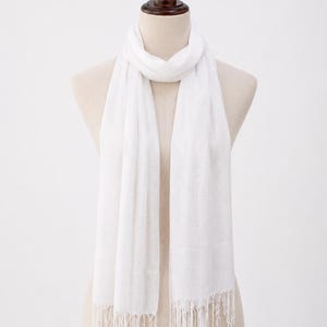 May include: A solid white rectangular scarf featuring a lightweight, semi-sheer fabric with a subtle woven texture. The scarf is draped around a mannequin neck and finished with delicate, thin fringe detailing along both bottom edges.