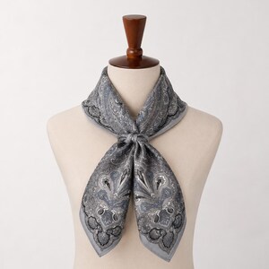 May include: A square scarf featuring a detailed paisley pattern in shades of grey, black, and white. The scarf is tied in a knot at the center, displaying intricate floral and teardrop motifs across the fabric, with a solid grey border edge.