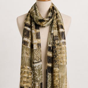 May include: A rectangular scarf featuring a patchwork design in shades of olive green, brown, black, and cream. The pattern consists of various geometric blocks, including squares, stripes, and leaf-like motifs, printed on a lightweight fabric.