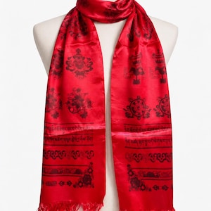 May include: A vibrant red scarf featuring a repeating pattern of dark maroon symbols, including wheels and floral motifs, alongside horizontal bands of Tibetan script. The scarf has a smooth, satin-like finish and is finished with fringed edges.
