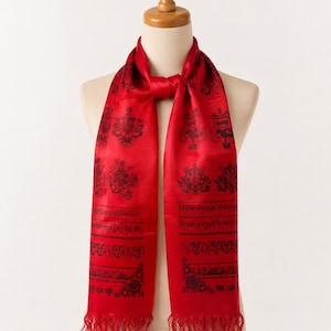 May include: A vibrant red scarf featuring a repeating black pattern of circular symbols, floral motifs, and lines of Tibetan script. The scarf has a smooth, satin-like finish and is finished with delicate red fringe along the bottom edges.