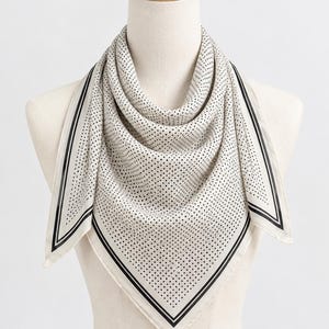 Op de afbeelding: Een vierkante sjaal in een gebroken witte kleur met een patroon van kleine zwarte stippen. De sjaal heeft een brede rand die wordt omlijnd door twee dunne zwarte lijnen langs de randen, wat een kader rond het stippenpatroon vormt.