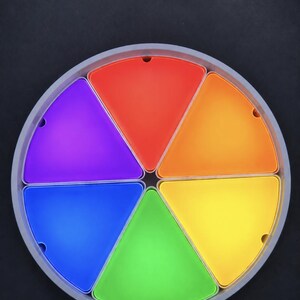 Könnte beinhalten: Ein kreisförmiges Tablett mit sechs herausnehmbaren dreieckigen Behältern in einem lebendigen Regenbogenspektrum aus Rot, Orange, Gelb, Grün, Blau und Violett. Jeder Behälter hat eine glatte Oberfläche und passt in den grauen Rahmen.