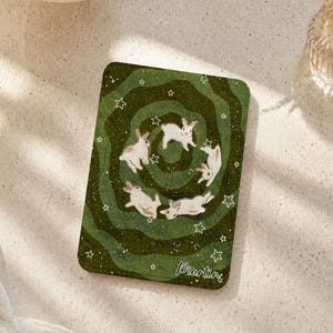 Puede incluir: Un posavasos rectangular verde con un patrón de vórtice en tonos verde oscuro y claro, cuatro conejos blancos y varios contornos de estrellas blancas. El nombre Martin está impreso en cursiva blanca en la esquina inferior derecha.