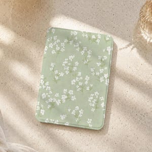 以下が含まれることがあります： 柔らかなセージグリーンの背景に、小さな白い花と細い緑の茎が繰り返される模様が施された長方形の電子書籍リーダーケース。ケースは角が丸くなっており、表面は滑らかなマット仕上げになっています。