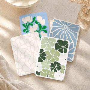 May include: Four rectangular coasters with rounded corners featuring floral patterns. One is light blue with white flowers and green stems, one is light blue with a white radial pattern, one is cream with white flowers, and one is green with white flowers.