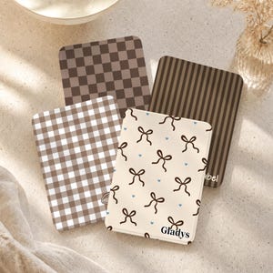 May include: Four rectangular keychains with rounded corners featuring distinct patterns: a brown checkerboard, a brown striped design, a brown and white gingham print, and a cream background with brown bows and small blue hearts labeled Gladys.