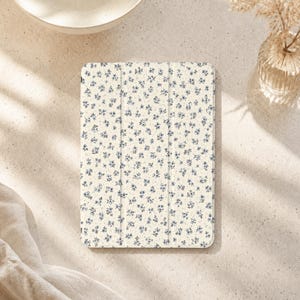 Puede incluir: Una funda rectangular para tableta con fondo color crema y un patrón denso y repetitivo de pequeñas flores de color azul oscuro. La funda presenta un diseño de tres pliegues con dos líneas verticales visibles en la parte frontal.