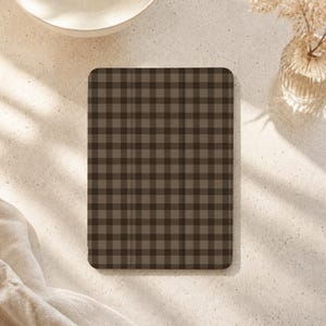 Könnte beinhalten: Eine rechteckige Tablethülle mit einem dunkelbraunen und hellbraunen Vichy-Karomuster. Die Hülle hat abgerundete Ecken und eine flache, glatte Oberfläche, die ein durchgehendes Gitterdesign auf der gesamten vorderen Außenfläche zeigt.