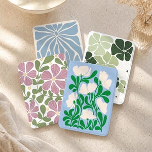 Puede incluir: Cuatro posavasos rectangulares con distintos patrones florales y abstractos. Uno tiene pétalos azules y blancos, otro muestra flores verdes y blancas, un tercero presenta flores rosas con hojas verdes y el cuarto tiene flores blancas sobre azul.