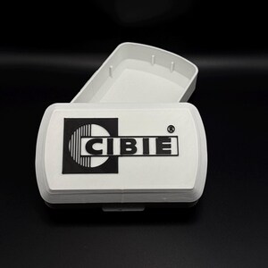 Peut inclure: Boîte rectangulaire blanche avec couvercle articulé, ouverte pour révéler l'intérieur. Le dessus de la boîte présente le logo noir et blanc "CIBIE". La boîte est posée sur un fond noir.