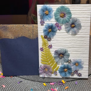 May include: A white greeting card with a horizontal ribbed texture, decorated with pressed blue flowers and a green fern leaf. Beside it sits a dark blue envelope featuring a textured gold band across the top edge, creating a contrasting set.