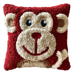 May include: A square throw cushion featuring a textured punch needle design of a monkey face. The monkey has deep red fur, a cream-coloured muzzle and ears, and black and white eyes. The design includes a red nose and a curved red mouth.