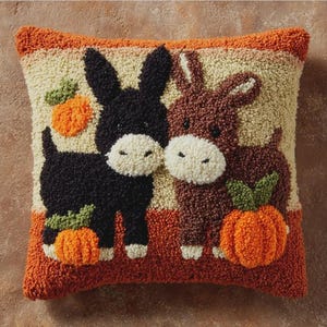 May include: A square throw pillow featuring a textured punch needle design of a black donkey and a brown donkey standing side by side. Each donkey is positioned next to a small orange pumpkin with green leaves against a cream background.