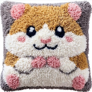 May include: A square tufted throw pillow featuring a stylized hamster design. The hamster has a golden-brown and white face, dark blue eyes, pink cheeks, and small pink paws. The background is a textured gray, and the entire surface is covered in dense, plush yarn loops.