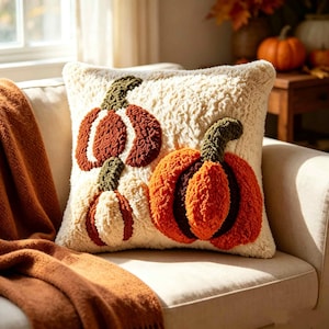 Pode incluir: Uma almofada quadrada com uma superfície texturizada e tufada na cor creme. A almofada apresenta três desenhos de abóboras bordadas em tons de laranja queimado, castanho escuro e creme, cada uma com um caule texturizado verde escuro.