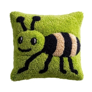 May include: A square throw pillow with a textured, tufted surface in a bright lime green color. The pillow features a large, stylized insect design in black and beige yarn, with two antennae, round eyes, a simple smile, and six thin legs.