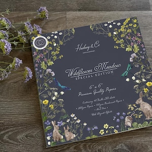 Könnte beinhalten: Ein quadratischer Papierblock mit dem Titel Wildflower Meadow Special Edition von Hackney & Co. Der dunkelgraue Einband zeigt Illustrationen von Wildblumen, Libellen und Hasen. Er misst 15,24 cm x 15,24 cm und enthält 40 beidseitig bedruckte Blätter.