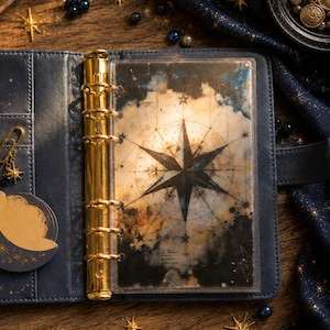 Peut inclure: Un classeur à anneaux en cuir bleu marine doté d'un mécanisme à six anneaux en métal doré. L'intérieur présente un intercalaire translucide imprimé d'un motif de boussole céleste sombre, avec une grande étoile, des lignes et des nuages.