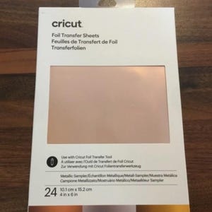 Può includere: Una confezione rettangolare bianca di fogli per trasferimento foil Cricut contenente 24 fogli da 10,1 cm per 15,2 cm. La confezione presenta un'ampia apertura rettangolare che rivela un foglio metallizzato color oro rosa e testo in varie lingue.